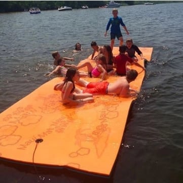 Children sitting on a large orange floating platform in the water with boats and a lifeguard in the background